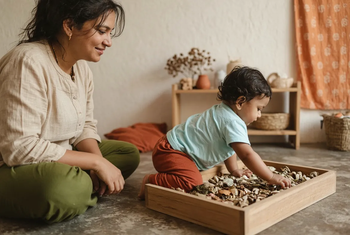 A toddler exploring a tray of natural materials near a seated adult